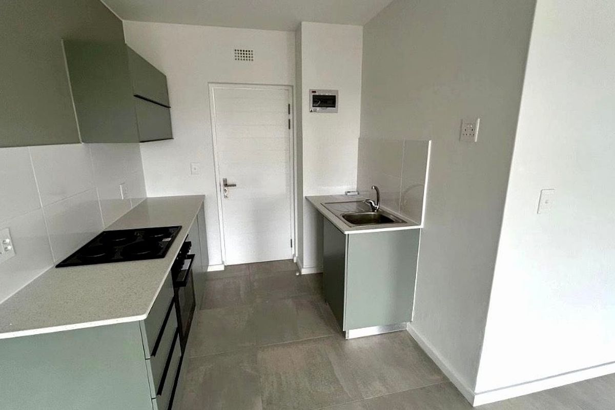A clean, modern kitchen featuring sage green cabinetry, stone countertops, integrated electric hob, oven, and stainless steel sink with neutral tiled flooring.