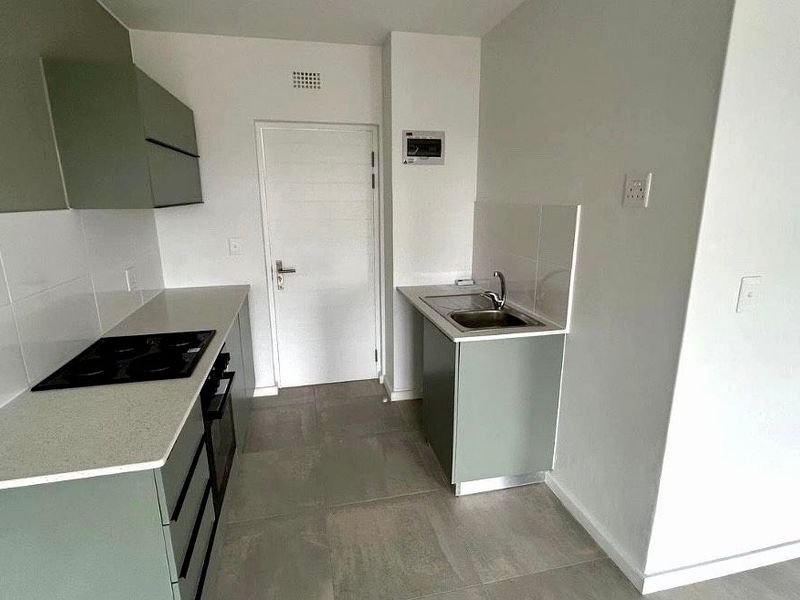 A clean, modern kitchen featuring sage green cabinetry, stone countertops, integrated electric hob, oven, and stainless steel sink with neutral tiled flooring.