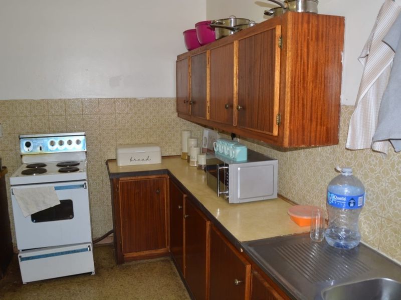 A dated kitchen featuring original wooden cabinetry, a laminate countertop, a stainless steel sink, and a freestanding stove with a tiled backsplash.