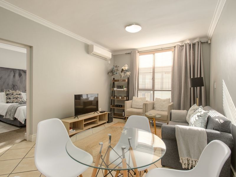 Open-plan living area featuring tiled flooring, crown molding, and a wall-mounted air conditioning unit, with an adjacent bedroom visible.