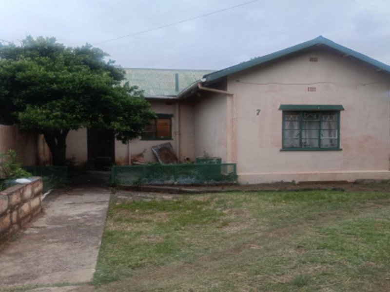Front view of a single-story house featuring a corrugated metal roof, concrete pathway, and stone retaining wall, showing signs of exterior wear.