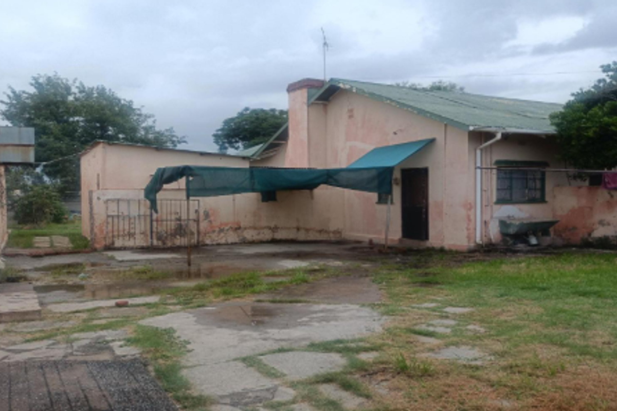 Rear view of the property showing a concrete patio area, weathered exterior walls, and a metal roof structure in need of maintenance.