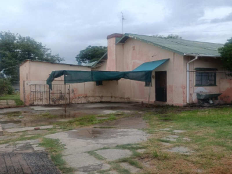 Rear view of the property showing a concrete patio area, weathered exterior walls, and a metal roof structure in need of maintenance.