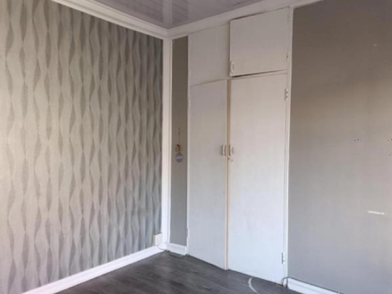 An empty bedroom featuring built-in cupboards, laminate flooring, and patterned wallpaper, offering a blank canvas for potential updates and interior design improvements.