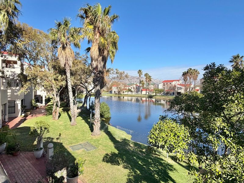 Scenic view overlooking a tranquil lake, landscaped garden area, and surrounding residential complex from an elevated balcony perspective.