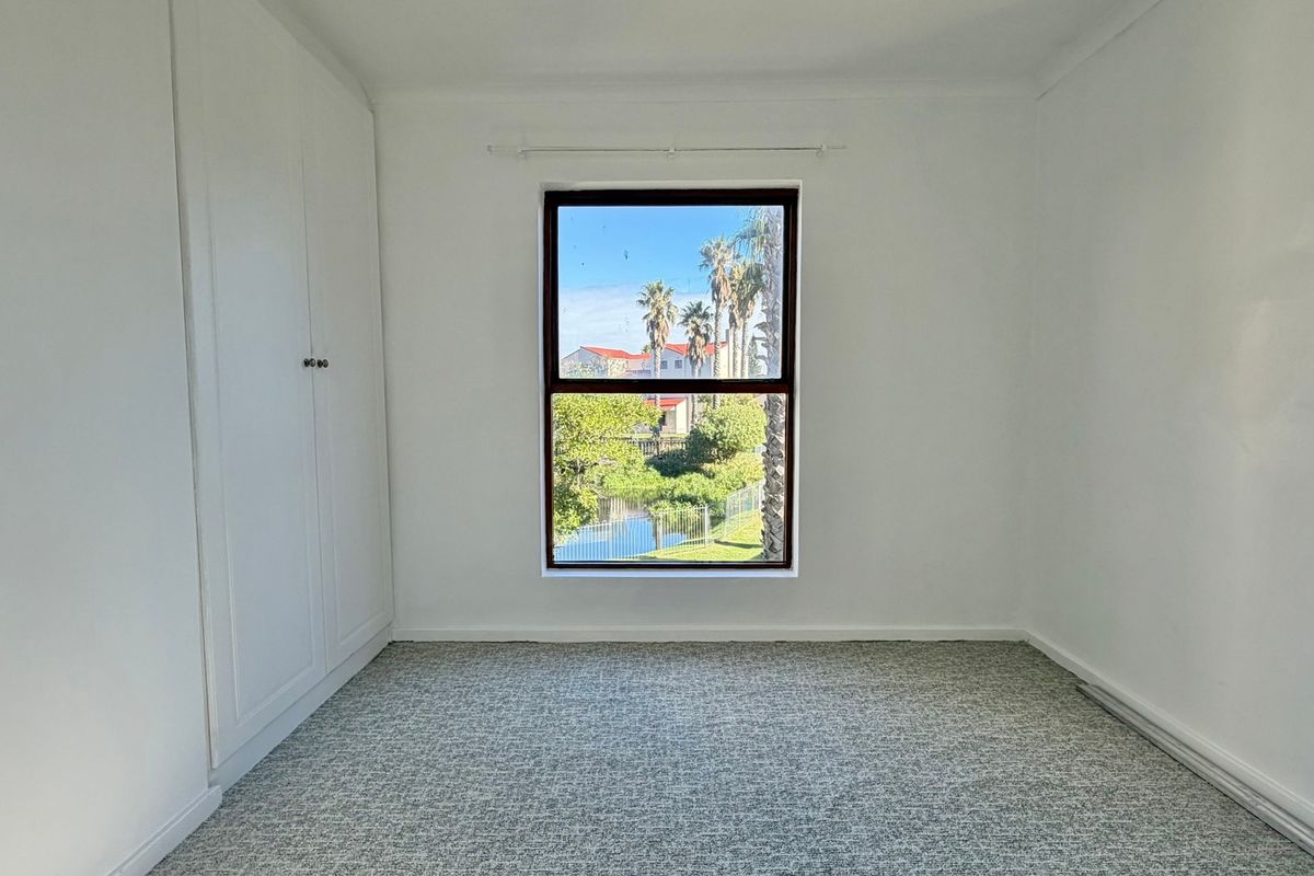 A bright, empty bedroom featuring built-in cupboards, carpeted flooring, and a window with a pleasant outdoor view.