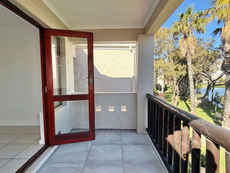 A covered balcony featuring tiled flooring and a wooden balustrade, offering pleasant views of the surrounding garden and water feature.