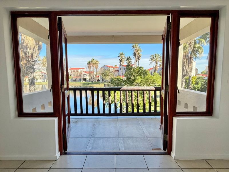 A bright, open balcony featuring tiled flooring and a wooden balustrade, offering scenic views of the surrounding estate and water feature.