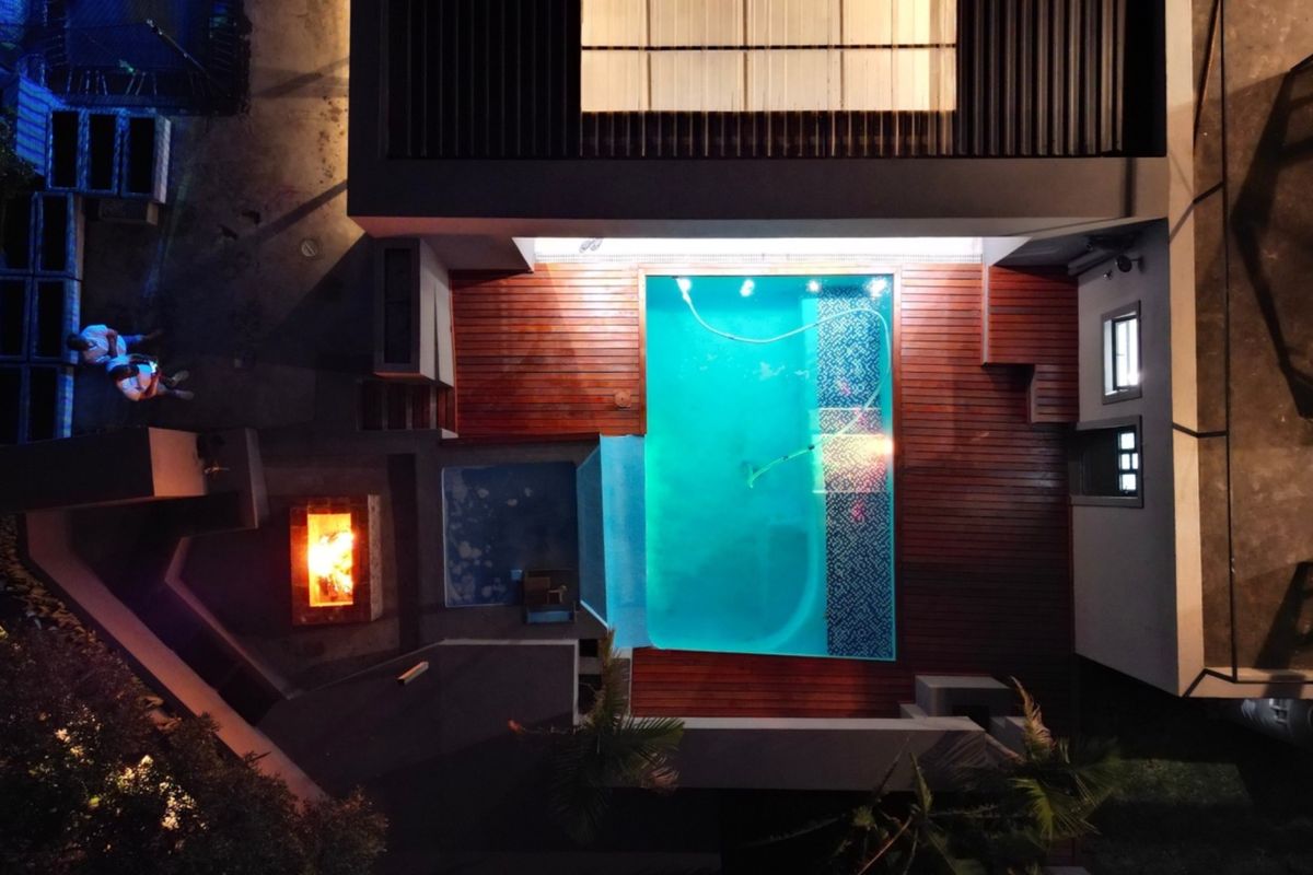 Aerial night view of a modern residential property featuring a swimming pool, timber decking, and a built-in fire pit area.