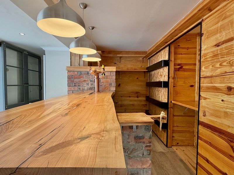 A custom-built indoor bar area featuring a natural timber countertop, exposed brick accents, and integrated wooden shelving with modern pendant lighting.
