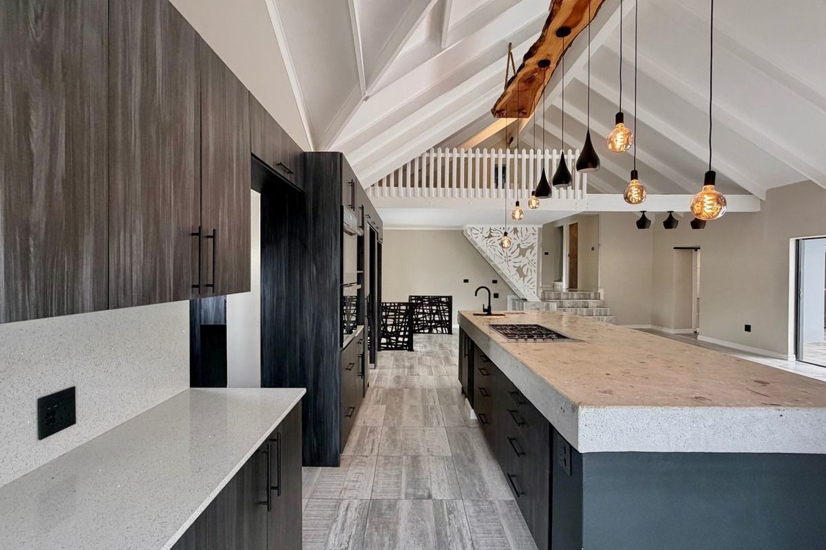 Modern open-plan kitchen featuring a large stone-topped island, dark cabinetry, exposed vaulted ceilings, and a mezzanine level overlooking the space.