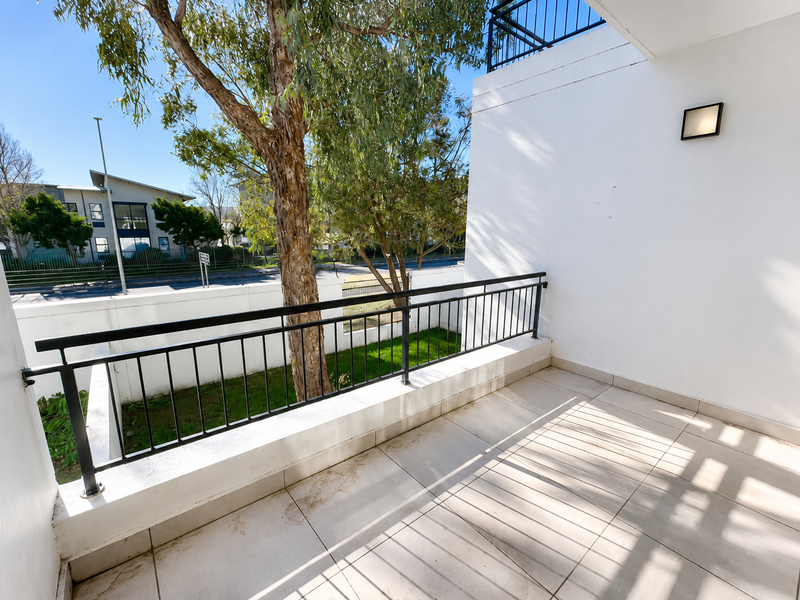 A private balcony featuring tiled flooring, a black metal balustrade, and an exterior wall light, overlooking a small garden area.