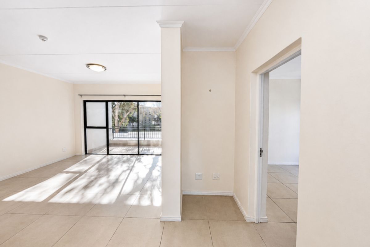 An empty, sunlit living area featuring neutral tiled flooring, coved ceilings, and a sliding glass door leading to an outdoor balcony.
