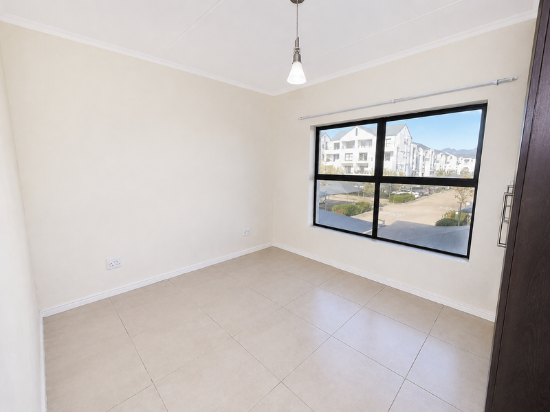 A bright, empty bedroom featuring neutral tiled flooring, a large aluminium-framed window, and a built-in cupboard, offering a clean and versatile living space.