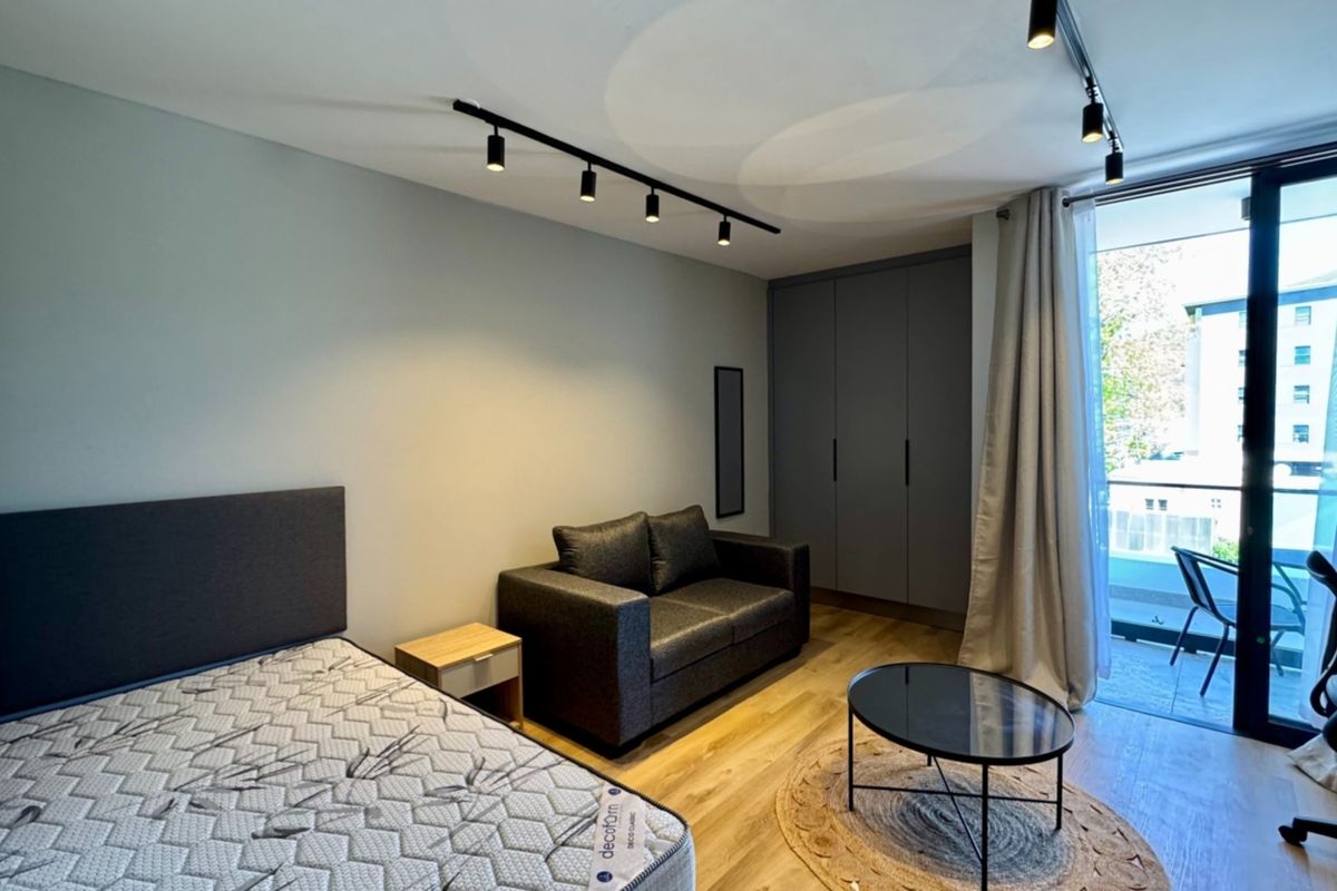 A modern bedroom featuring built-in wardrobes, track lighting, and direct access to a private balcony through a large sliding glass door.