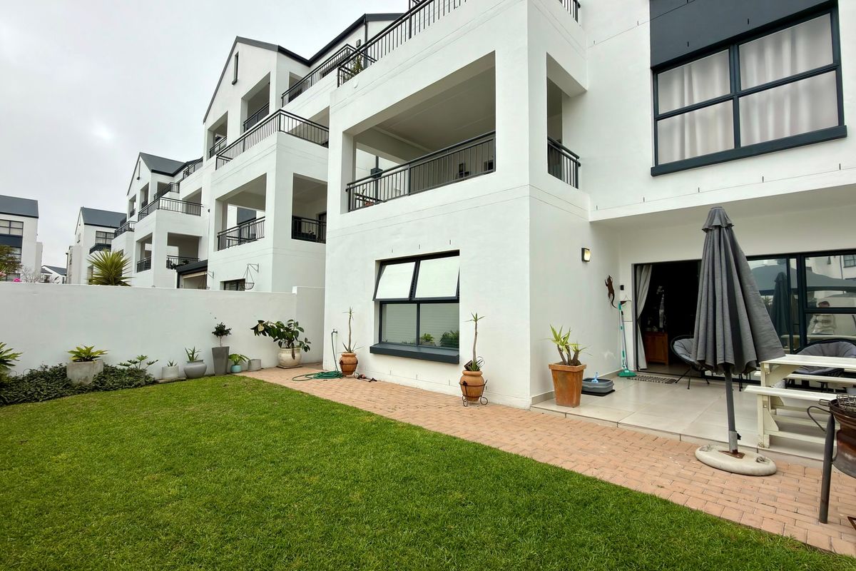 Rear view of the apartment complex featuring a private garden, brick-paved walkway, tiled patio area, and upper-level balconies with modern finishes.