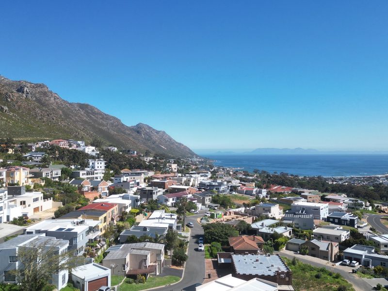 An elevated aerial view showcasing the coastal residential neighbourhood, surrounding mountain range, and ocean horizon.