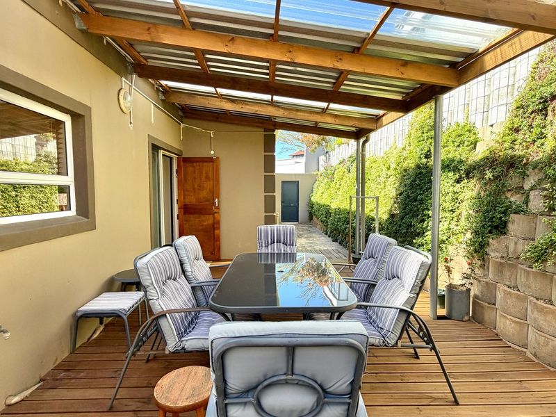 A covered outdoor patio area featuring timber decking, a pergola roof structure, and a stone retaining wall, providing a functional space for outdoor dining.