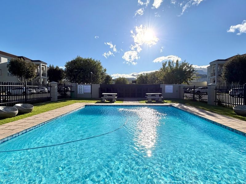 A well-maintained communal swimming pool area featuring paved surrounds, manicured lawn, and secure metal perimeter fencing within an apartment complex.