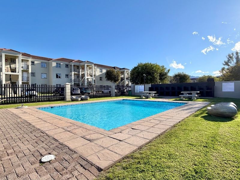 Communal swimming pool area featuring a paved deck, manicured lawn, and built-in braai facilities within a secure apartment complex.