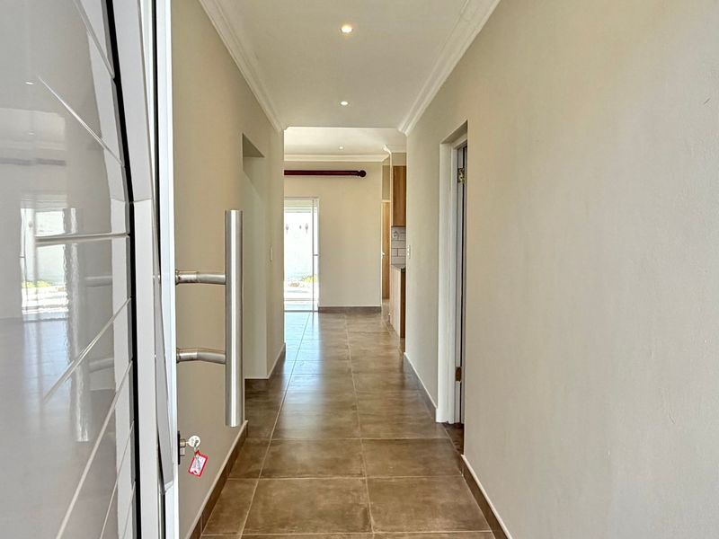 A bright, neutral-toned hallway featuring tiled flooring, recessed downlighting, and clean architectural lines leading towards the living area.