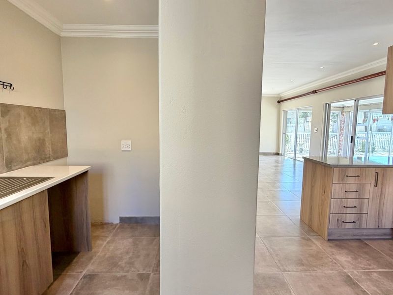 Open plan kitchen and scullery area featuring modern wood-finish cabinetry, stone countertops, tiled flooring, and direct access to the outdoor patio through sliding glass doors.