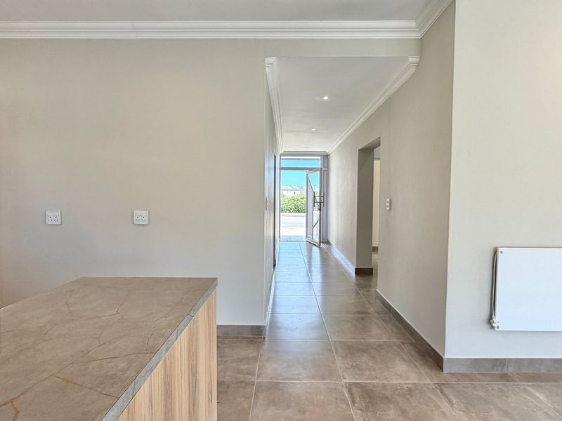 A bright, neutral hallway featuring tiled flooring, coving, and built-in shelving, leading towards an exterior doorway with natural light.