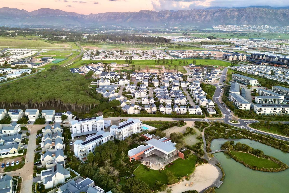 An aerial view of a residential estate featuring modern housing, solar installations, landscaped green spaces, and a scenic lake with mountain views.