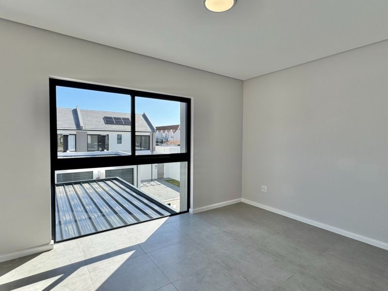 A bright, modern, and empty bedroom featuring neutral grey floor tiles, clean walls, and a large window providing natural light and a view of the complex.