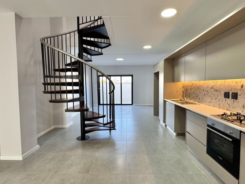 Modern open-plan living area featuring a striking spiral staircase, sleek cabinetry, integrated kitchen appliances, and neutral tiled flooring leading to a rear patio.