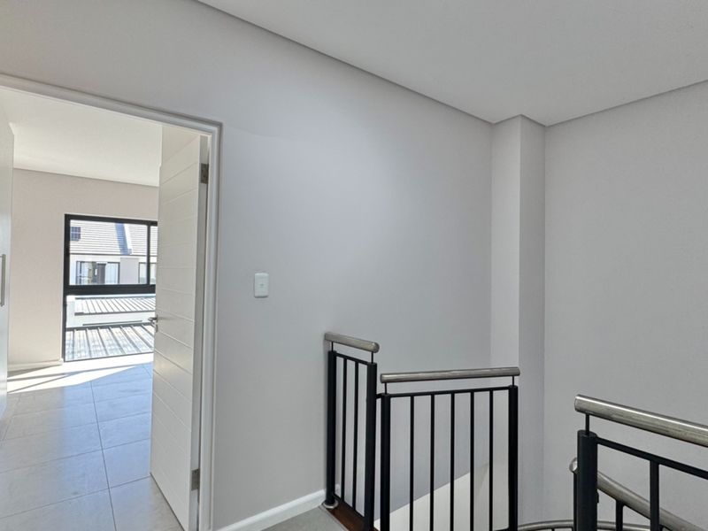 A bright, modern hallway featuring a sleek metal balustrade and tiled flooring, leading into an adjacent room with natural light.