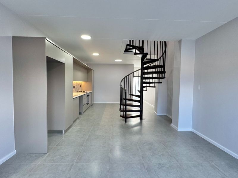 Modern open-plan living area featuring a striking black spiral staircase, sleek built-in kitchen cabinetry, and neutral tiled flooring throughout.