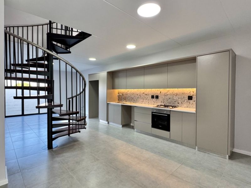 Modern open-plan kitchen featuring sleek cabinetry, terrazzo backsplash, integrated appliances, and a striking spiral staircase, set against neutral tiled flooring.