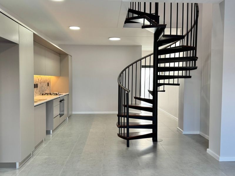 Modern open-plan living area featuring a striking spiral staircase, sleek built-in kitchen cabinetry, stone backsplash, and contemporary tiled flooring throughout.