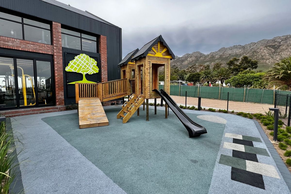 A well-maintained outdoor play area featuring a wooden jungle gym with a slide, set on modern rubberized flooring adjacent to a brick building.