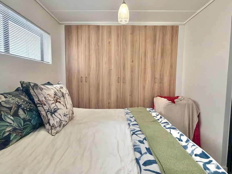 A well-maintained bedroom featuring floor-to-ceiling wood-finish built-in wardrobes, a horizontal window with venetian blinds, and a central pendant light fixture.