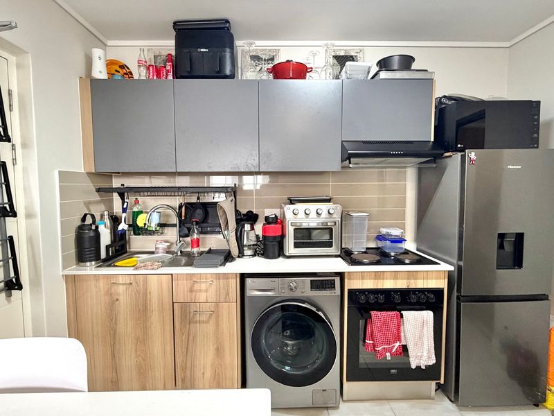 Compact kitchen featuring modern wood-grain lower cabinetry, grey upper cabinets, tiled backsplash, built-in oven, hob, extractor fan, and an integrated washing machine.