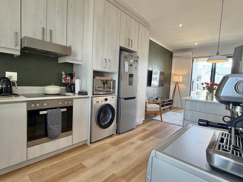 Modern open-plan kitchen and living area featuring light wood-look cabinetry, integrated appliances, laminate flooring, and direct access to a balcony.