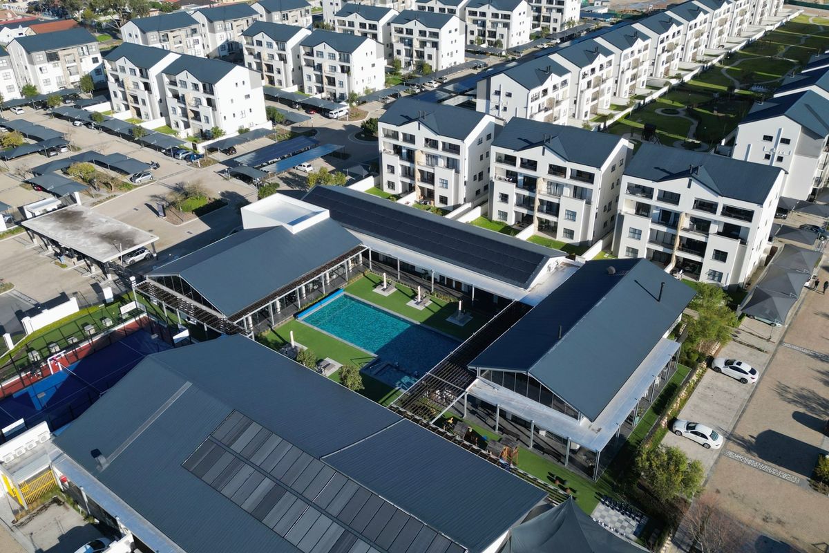 Aerial view of a modern apartment complex featuring a central swimming pool, sports facilities, solar panels, and ample parking within a landscaped estate.