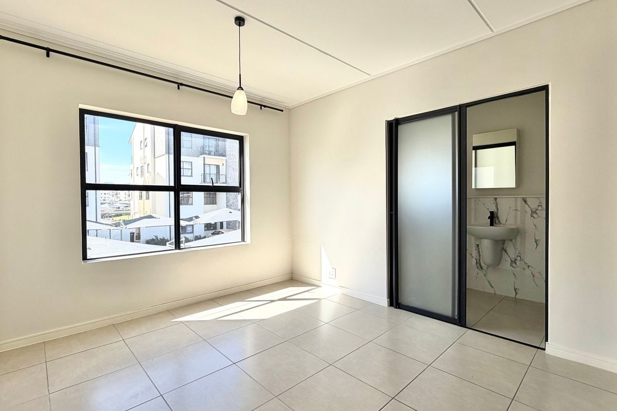 A bright, modern bedroom featuring neutral tiled flooring, a large window, and an integrated en-suite bathroom with a sleek sliding glass door.
