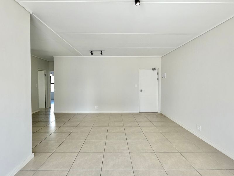 A spacious, empty living area featuring neutral tiled flooring, clean white walls, and modern track lighting, offering a versatile blank canvas.