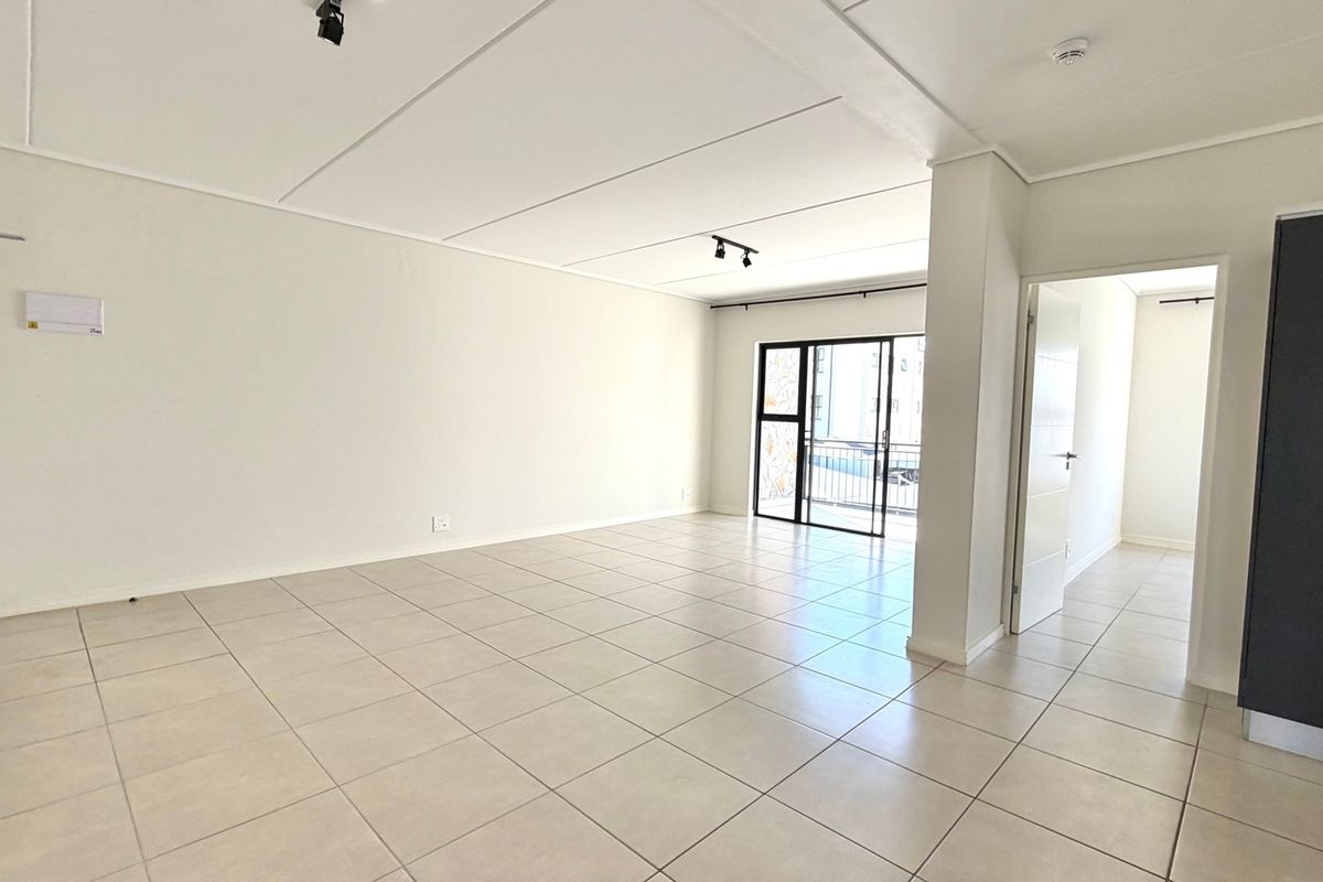 A bright, spacious, and empty living area featuring neutral tiled flooring, modern track lighting, and a sliding glass door leading to an outdoor space.