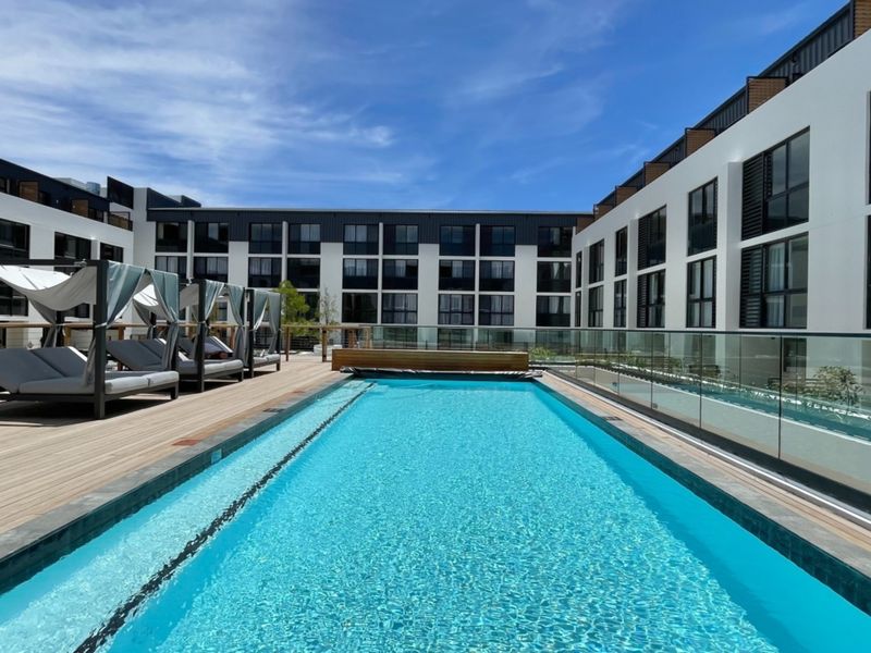 A modern apartment complex featuring a pristine swimming pool with timber decking and glass balustrades, set within a contemporary residential courtyard.