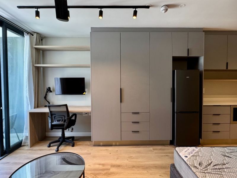 Modern open-plan studio apartment featuring integrated desk space, extensive built-in cabinetry, track lighting, and direct access to a private balcony through sliding glass doors.