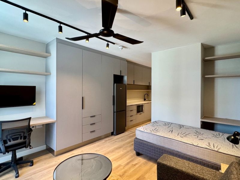 Modern open-plan studio apartment featuring a sleek kitchen, built-in cabinetry, integrated desk space, and contemporary track lighting with wood-look flooring.