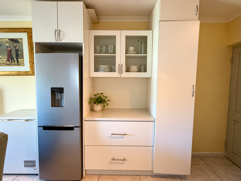 A functional kitchen storage unit featuring white cabinetry, a glass-fronted display section, and a dedicated space for a refrigerator.