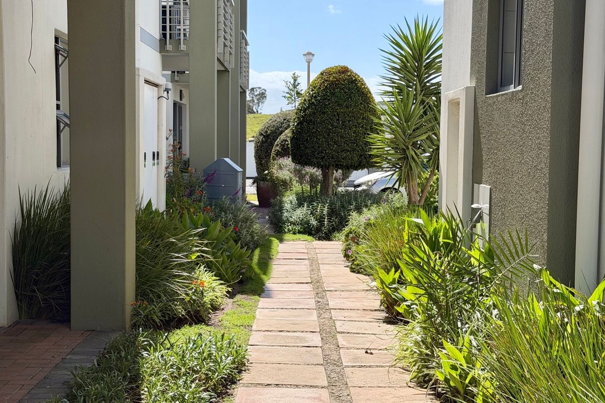 A well-maintained paved walkway leading through a landscaped garden between apartment building blocks, featuring established greenery and clean exterior finishes.