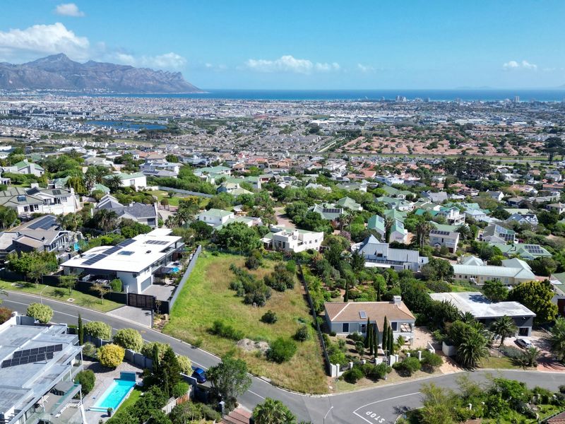 An aerial view of a vacant residential plot situated within an established suburban neighborhood, featuring expansive views of the surrounding landscape and coastline.