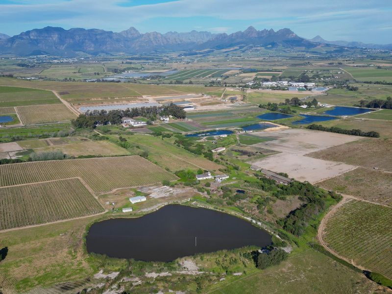 An expansive aerial view of a large farm property featuring vineyards, agricultural fields, and a prominent farm dam set against a mountain backdrop.