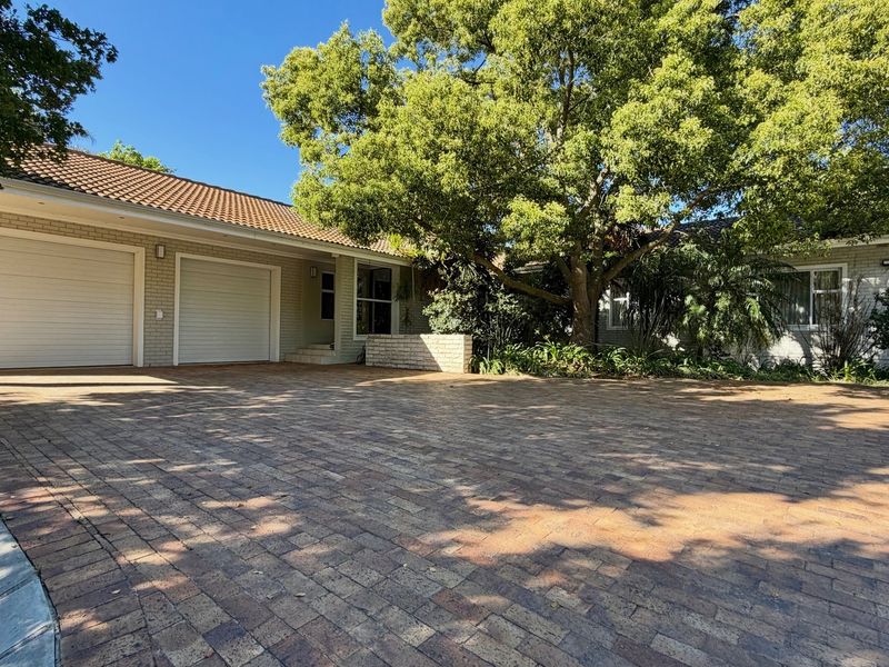 Front exterior view of the property featuring a spacious brick-paved driveway, a double garage, and established mature trees providing shade.